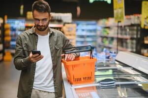 a el supermercado hermoso hombre usos teléfono inteligente y mira a nutricional valor de el Enlatado bienes. él es en pie con compras carro en Enlatado bienes sección. foto