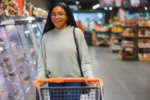 African american woman with shopping cart trolley in the supermarket store look on mobile phone photo