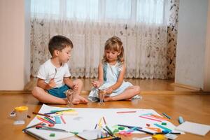 Kids drawing on floor on paper. Preschool boy and girl play on floor with educational toys - blocks, train, railroad, plane. Toys for preschool and kindergarten. Children at home or daycare. photo
