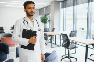 Portrait of male indian doctor wearing white coat having open door on clinic corridor as background photo