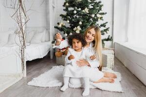 Merry Christmas and Happy Holidays Cheerful mom and her cute daughter at Christmas tree. Parent and little child having fun near Christmas tree indoors. Loving family with presents in room. photo