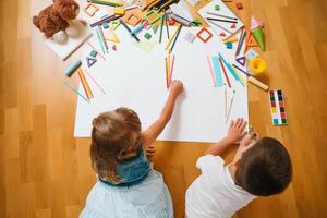 Kids drawing on floor on paper. Preschool boy and girl play on floor with educational toys - blocks, train, railroad, plane. Toys for preschool and kindergarten. Children at home or daycare. Top view. photo