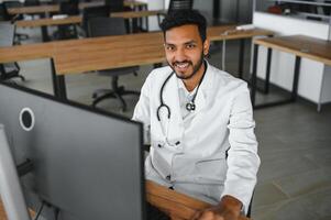 People, occupation and medicine concept. Smiling male Indian Arab doctor in white coat, sitting in medical office at desk with laptop photo