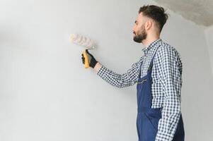 joven sonriente profesional trabajador en azul uniforme en pie con pintar rodillo en nuevo Departamento para reparando terminado gris paredes fondo, Copiar espacio. foto