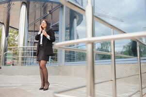 Business woman with coffe and talking on the phone near office. Young woman with smartphone standing against street blurred building background. Business photo of beautiful girl in casual suite