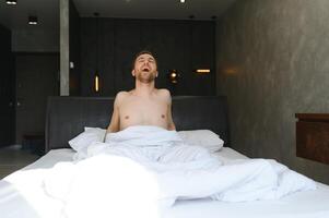 joven hombre despertar arriba y subir manos extensión en blanco cama en el Mañana con luz de sol mediante ventana foto