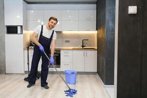 Man as a professional cleaner in blue uniform washing floor with mopping stick and bucket in the living room of the apartment photo