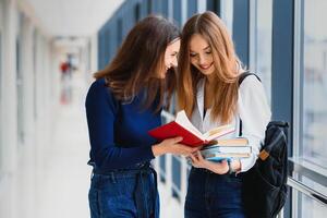 Two young female students standing with books and bags in the hallway University speaking each other. photo