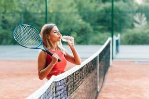 Female tennis player with towel on her shoulders drinking water after match. photo