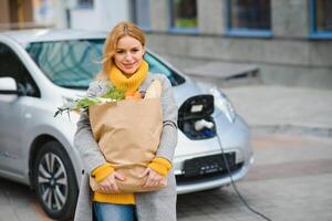 Charging electro car at the electric gas station. Woman standing by the car. Lady with foodstuff photo