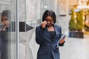 Confident businesswoman outside. Black woman in office suit standing near urban glass wall and using mobile phone. African American business woman concept photo