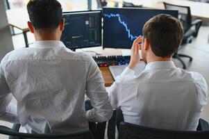 Two successful trader sitting in office and looking at screen, checking cryptocurrency information data on finance market graph, pointing on monitor photo