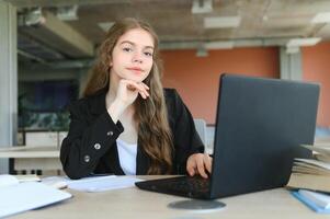 A beautiful caucasian female student is studying in college remotely. She is sitting with a laptop and a notepad and concentrated is watching a video conference lesson photo