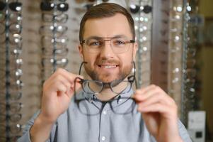 Big choice. Stylish man with beard choosing glasses in the optics store. photo