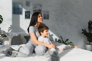 Young mother with her 5 years old little son dressed in pajamas are relaxing and playing in the bed at the weekend together, lazy morning, warm and cozy scene. Family home shoot. photo