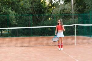 niño jugando tenis en al aire libre corte. pequeño niña con tenis raqueta y pelota en deporte club. activo ejercicio para niños. verano ocupaciones para niños. formación para joven niño. niño aprendizaje a jugar foto