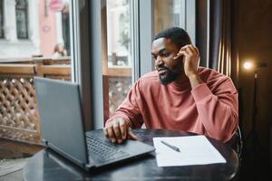 afro americano negociaciones vídeo charla en computadora se sienta en café con taza de café. negro hombre llamadas en ordenador portátil en vídeo enlace y habla él usa en camisa y traje chaqueta. vídeo enlace en línea conexión foto