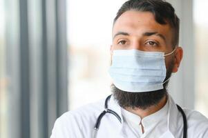 Male pensive thoughtful arab man therapist surgeon wear white uniform medical face mask standing at workplace looking out window preventing spread infection pandemic outbreak close-up doctor portrait. photo