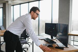 Man trader in formalwear sitting at desk in frot of monitors with charts and data at office browsing laptop checking documents analyzing stocks price changes concentrated. photo