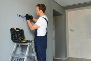 Electrician in uniform mounting electric sockets on the white wall indoors photo