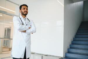 Portrait of male indian doctor wearing white coat having open door on clinic corridor as background photo