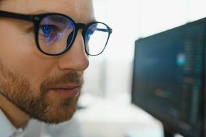 Young business man trader wearing glasses looking at computer screen with trading charts reflecting in eyeglasses watching stock trading market financial data growth concept, close up. photo