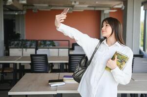 retrato de un contento alegre sonriente joven estudiante Chica de escuela dama con largo pelo en pie en vacío salón de clases mirando cámara tomar un selfie foto