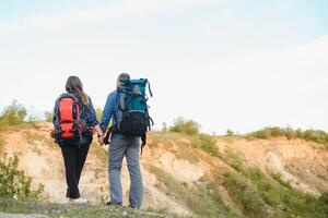 espalda ver de joven par mochileros con grande mochilas participación manos y caminando a lo largo un la carretera con hermosa montaña paisaje en antecedentes. foto