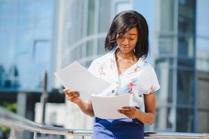 Content formal African American woman in suit standing on sidewalk on street and reading papers in file against contemporary building photo