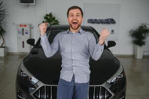 Emotional handsome guy raising hand up and smiling at camera. Rich man buying auto at luxury dealership salon photo