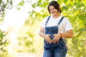 retrato de un contento y orgulloso embarazada mujer mirando a su barriga en un parque a amanecer con un calentar espalda ligero en el antecedentes foto