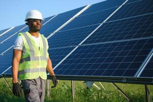 Competent energy engineer in grey overalls and white helmet checking solar panels while walking on field. African american man carrying clipboard and container with instruments photo