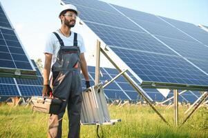 Male arabian engineer in helmet and brown overalls checking resistance in solar panels outdoors. Indian man working on station. photo