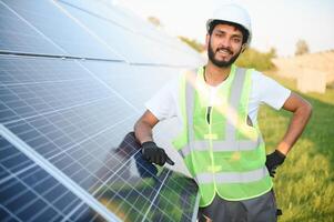 Male arabian engineer in helmet and brown overalls checking resistance in solar panels outdoors. Indian man working on station. photo
