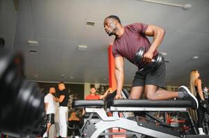 Young African American man sitting and lifting a dumbbell close to the rack at gym. Male weight training person doing a biceps curl in fitness center photo