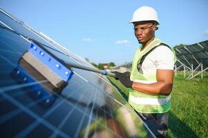 Alternative power plant worker in uniform cleaning solar panels with mop. Handsome African-American taking care of equipment. photo