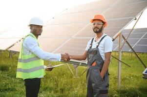 Group of multi ethnic people in safety helmets standing at solar station. Two engineers and technician examining plan of panels outdoors. photo