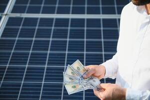 male hand hold hundred dollar at the background of solar station. concept of generation photo
