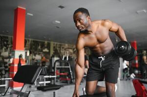 Young African American man sitting and lifting a dumbbell close to the rack at gym. Male weight training person doing a biceps curl in fitness center photo