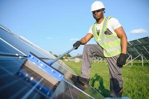 Alternative power plant worker in uniform cleaning solar panels with mop. Handsome African-American taking care of equipment. photo