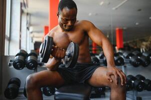 Young African American man sitting and lifting a dumbbell close to the rack at gym. Male weight training person doing a biceps curl in fitness center photo