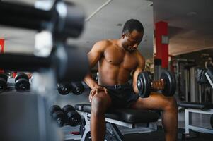 Young African American man sitting and lifting a dumbbell close to the rack at gym. Male weight training person doing a biceps curl in fitness center photo