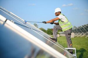 alternativa poder planta trabajador en uniforme limpieza solar paneles con fregar. hermoso afroamericano tomando cuidado de equipo. foto