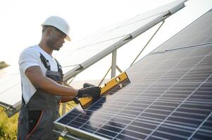 retrato de joven hermoso africano americano artesano en protector casco. hombre en uniforme y con herramientas en pie entre solar paneles foto