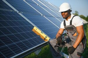 African american man in safety helmet and glasses tighten nuts on solar panels with screwdriver. Competent technician using tools while performing service work on station photo