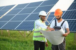 grupo de multi étnico personas en la seguridad cascos en pie a solar estación. dos ingenieros y técnico examinando plan de paneles al aire libre. foto