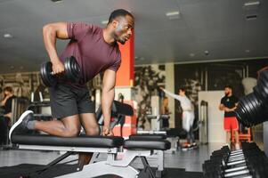 Hot african american young man bodybuilder lifting barbell at gym, working on his arms, looking at copy space. Black muscular shirtless guy having biceps workout session. Healthy lifestyle concept. photo