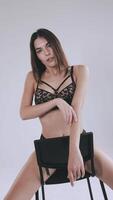 un profesional modelo en negro ropa interior en un blanco antecedentes poses en un negro silla. hermosa niña en lencería. sexy mujer coquetea en frente de el cámara en el estudio. video