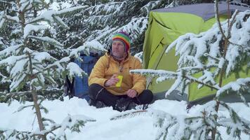 resande nära en tält i de skog i vinter. en man är vilar och dricka te från en gul mugg. resa begrepp. 4k video