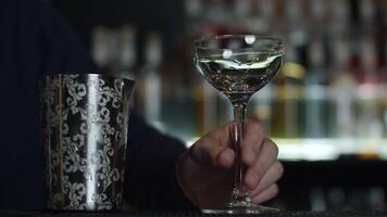 Close up of a bartender putting ice cubes into the small empty cocktail glass. Media. Details of making a drink with blurred bottles of alcohol on the background. video